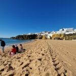 Vista panoramica della grande e deserta spiaggia di Albufeira, in Algarve, a Gennaio. Poche persone sedute in abiti invernali godono del sole e della vista del mare nella sua interezza, evidenziando la tranquillità e l'ampiezza dello spazio in bassa stagione.
