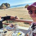 Coppia (Ilaria e Gabriel di Tripilare) seduta al tavolo all'aperto, vestita con maglioni leggeri e occhiali da sole, che pranza in una giornata di sole in Algarve a Gennaio. L'immagine evidenzia il clima mite e il piacere di stare fuori in pieno inverno.