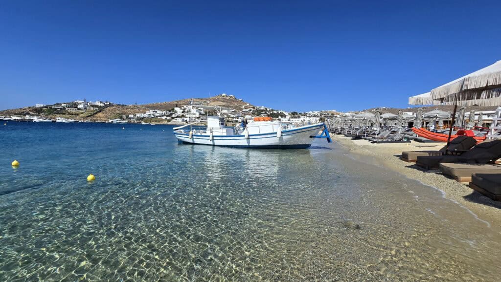 Spiaggia di Ornos Mykonos: Guida Completa, Consigli e Prezzi 2 Spiaggia di Ornos Mykonos: barca ancorata nell'acqua cristallina, con i lettini posti così a riva che le onde arrivano quasi a toccarli.