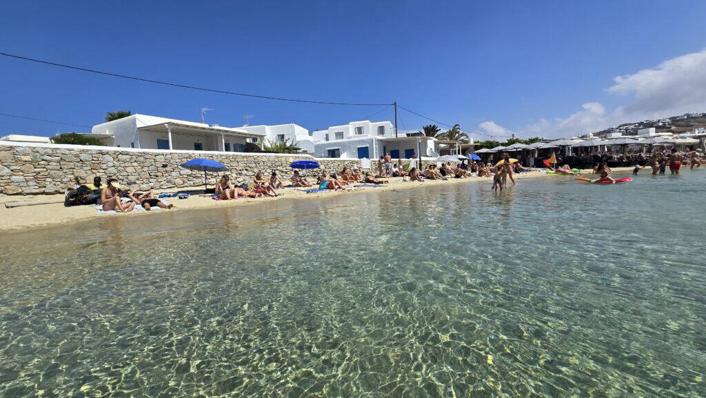 Spiaggia di Ornos Mykonos: Guida Completa, Consigli e Prezzi 4 Area limitata di spiaggia libera a Ornos Beach Mykonos, con acqua cristallina e bagnanti che prendono il sole vicino alle case bianche dell'isola.