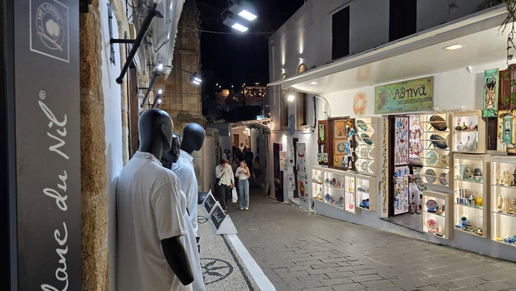 Via principale di Lindos (Rodi) di sera, illuminata, con persone che passeggiano e boutique aperte. Illustra l'atmosfera vivace del centro storico.