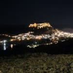 Panorama notturno di Lindos Rodi con le case bianche illuminate e l'imponente Acropoli che brilla sulla sommità.